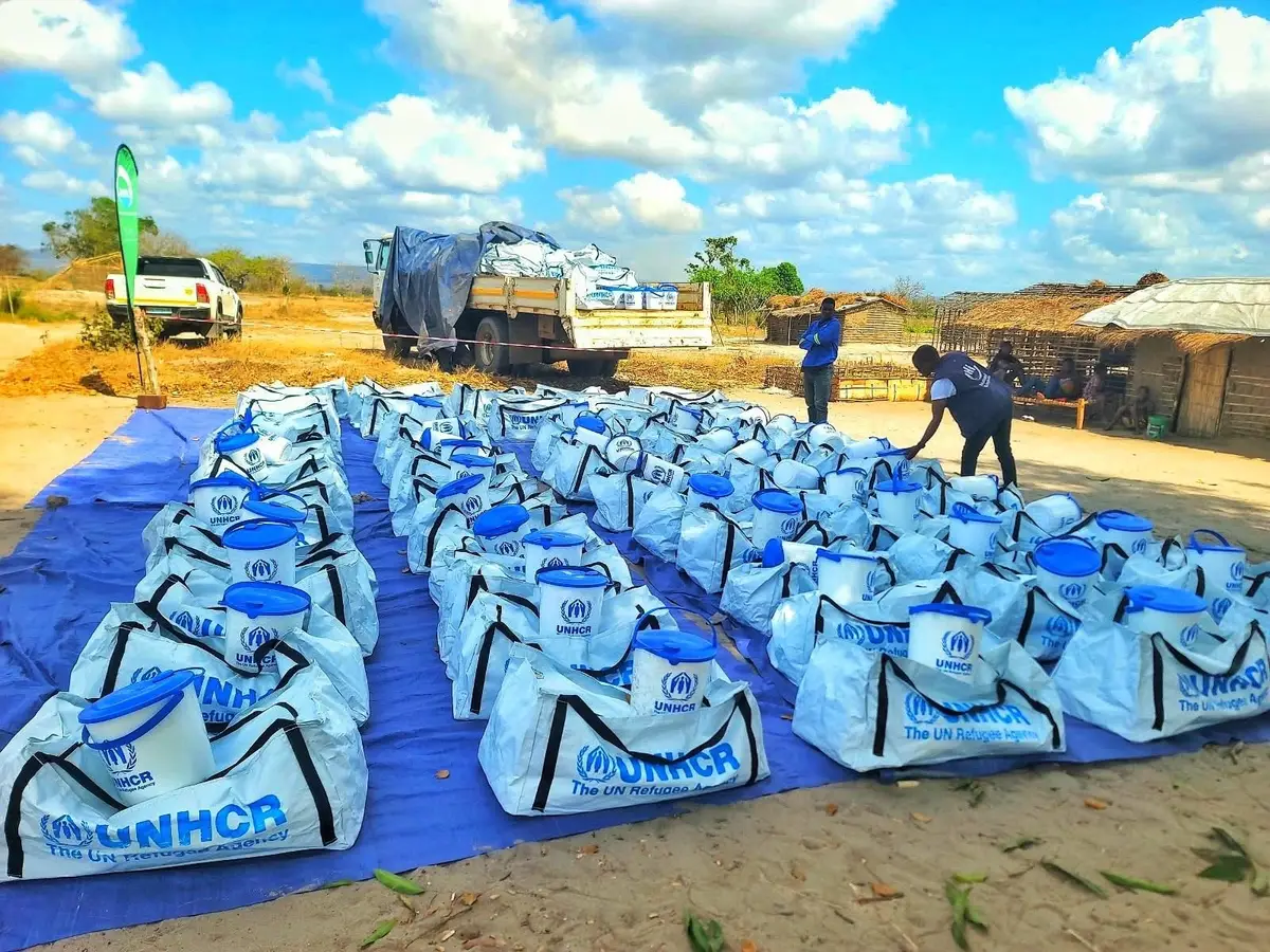 Kits do ACNUR alinhados sobre lonas, prontos para distribuição, com um camião ao fundo